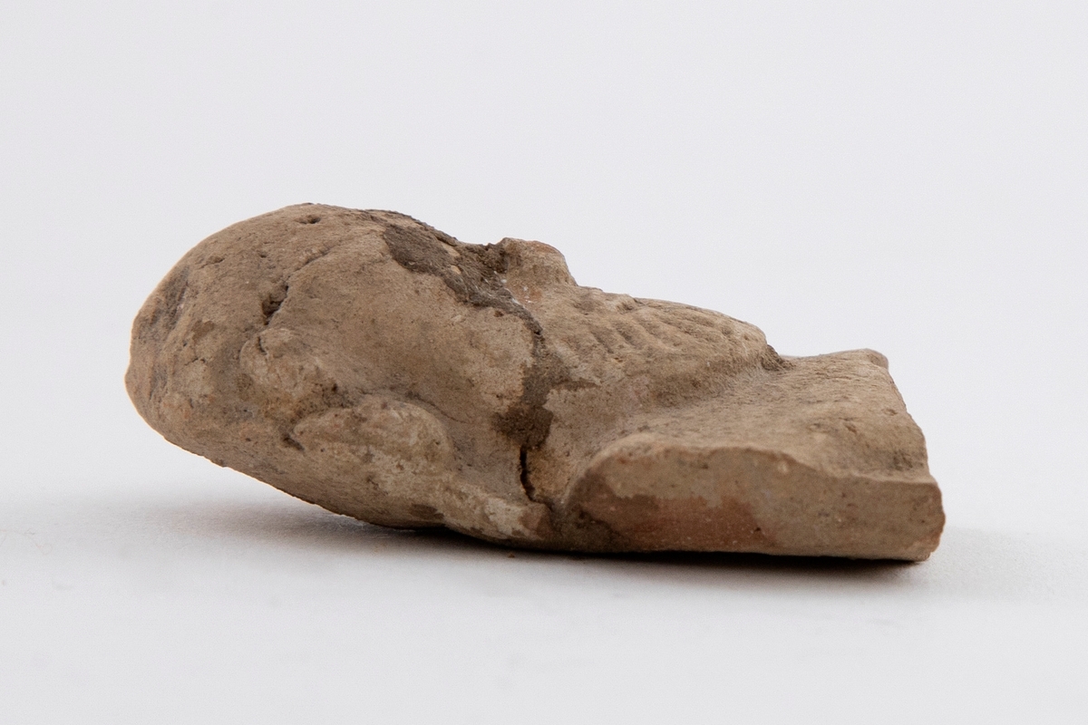 Fragment av figurin i terrakotta, øvre del av kroppen er bevart. Fremstiller en faun med skjegg. Synlige brudd og rester av jord.