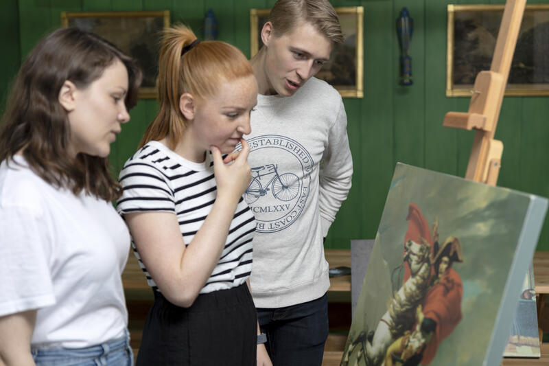 Halvnært bilde av tre videregåendeelever som står og ser på en kopi av et berømt maleri av Napoleon til hest i Grønnstua i Eidsvollsbygningen