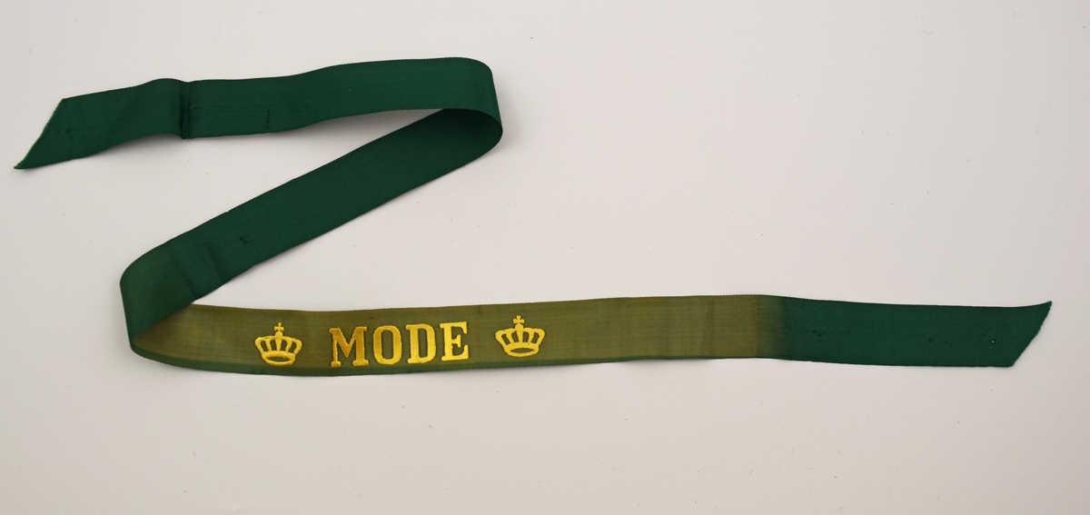 Mössband av grönt sidenrips. Guldfärgad text, "Mode", med två kronor på vardera sida. Blekt i färgen.