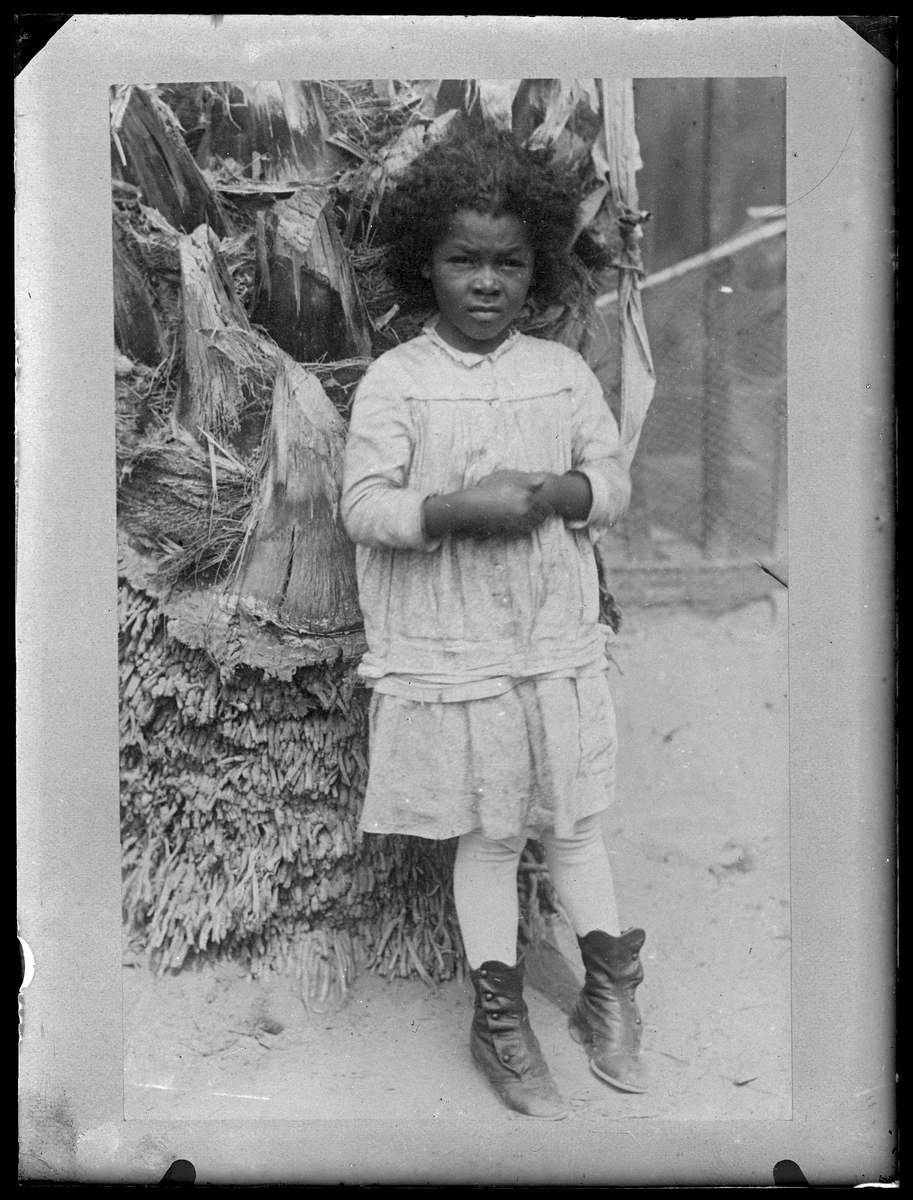 Bilden föreställer en liten flicka i västerländska kläder och skor som lutar sig mot en palm.
