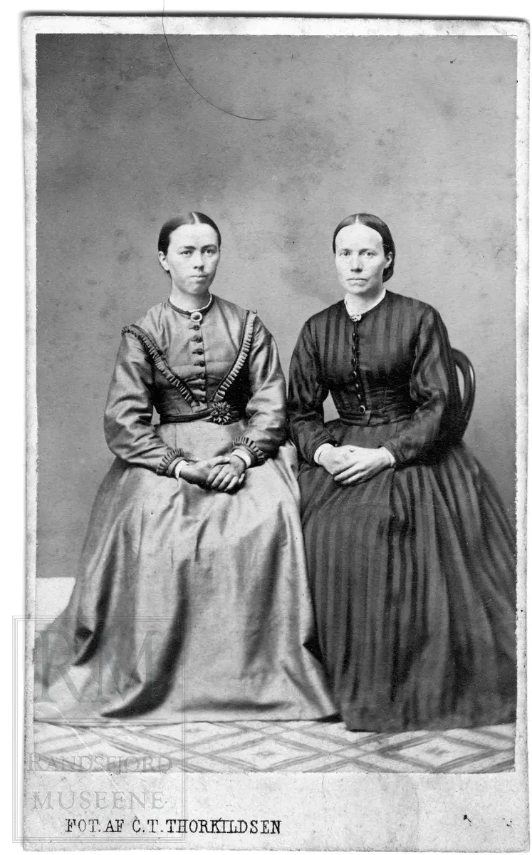 To kvinner sitter i hver sin stol. Begge har vide kjoler med knepping i front og brosje i halsen.
