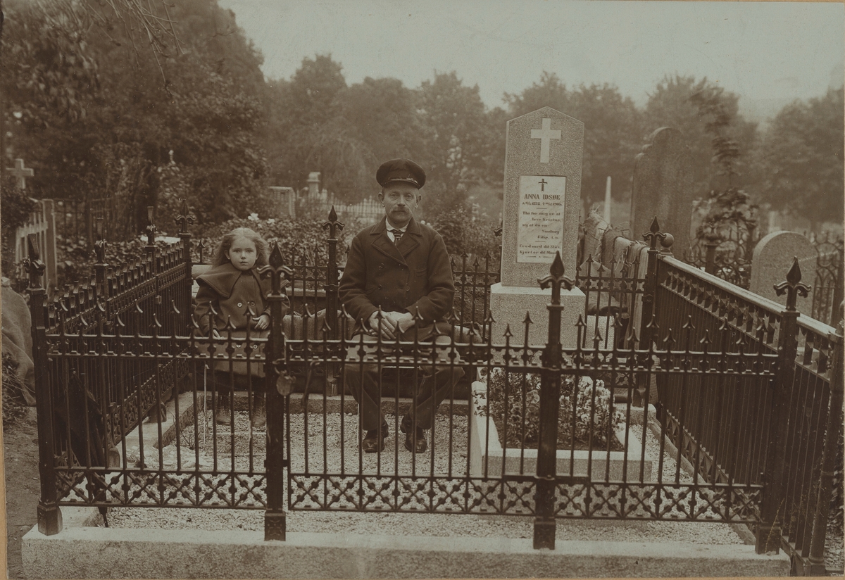 Viser gravstedet til Anna Odsøe (1878-1906). Gravstedet er inngjerdet med smijernsgjerde. En mann og en liten pike sitter på klappstoler.