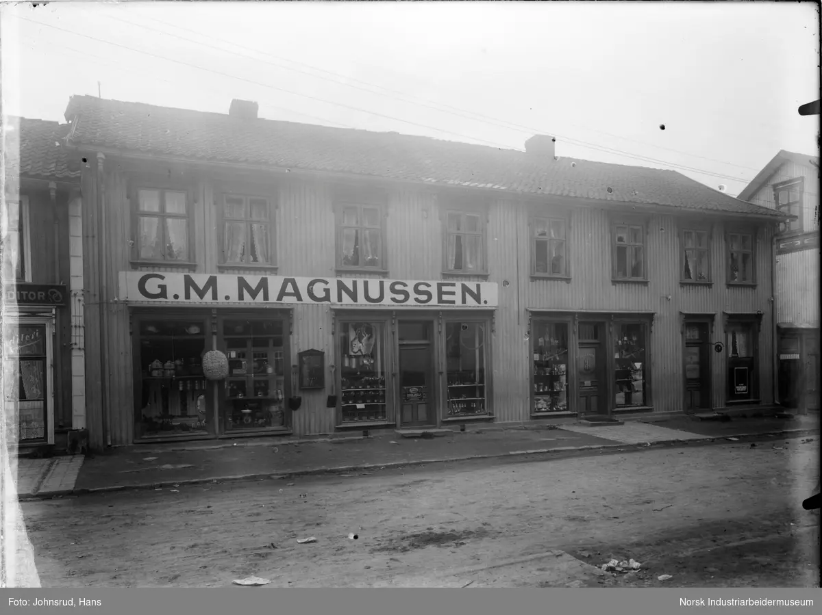 Eksteriør av G.M. Magnussens butikk i kongsberg. Toetasjers trebygg der det er utstillinger i alle vinduer i første etasje. Bygning med konditori delvis synlig til venstre.