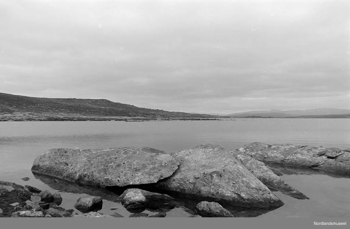 Fjord, fjell og fjæresteinene tatt fra Nasa, Silbjaure. Fjæra i forgrunnen. Fjellene i bakgrunnen. Overskyet.