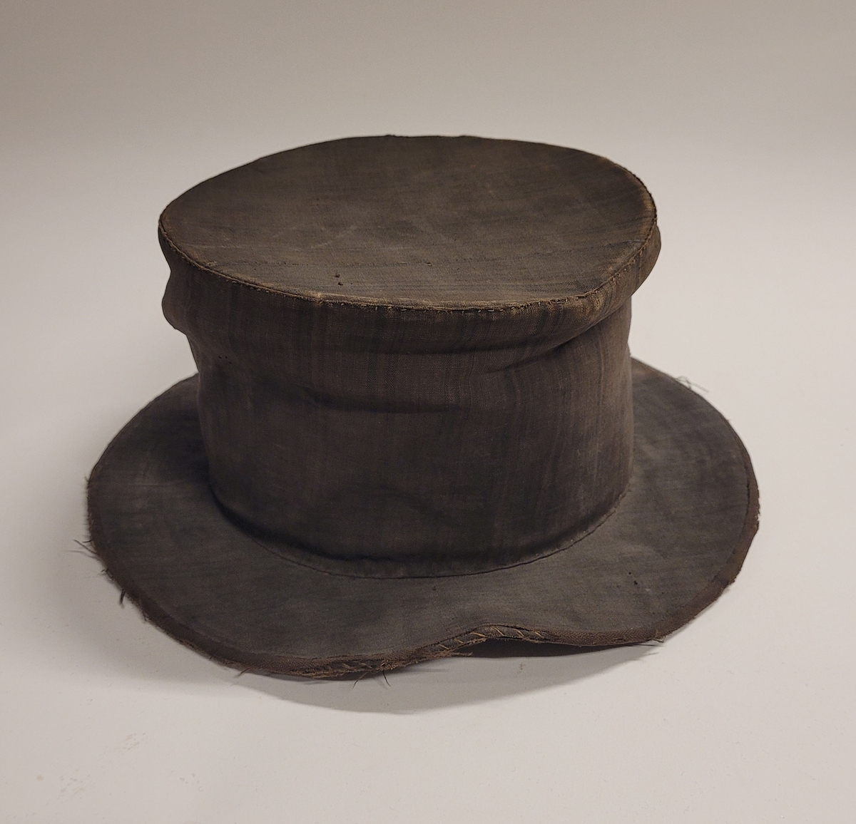 Hög hatt med brätten, överdragen med mörkt randigt linnetyg i fyra färger, övervägande brunt. Svettrem längs insidan av svart läder. Fodrad med vit lärft. Skullen uppbyggd av halm.