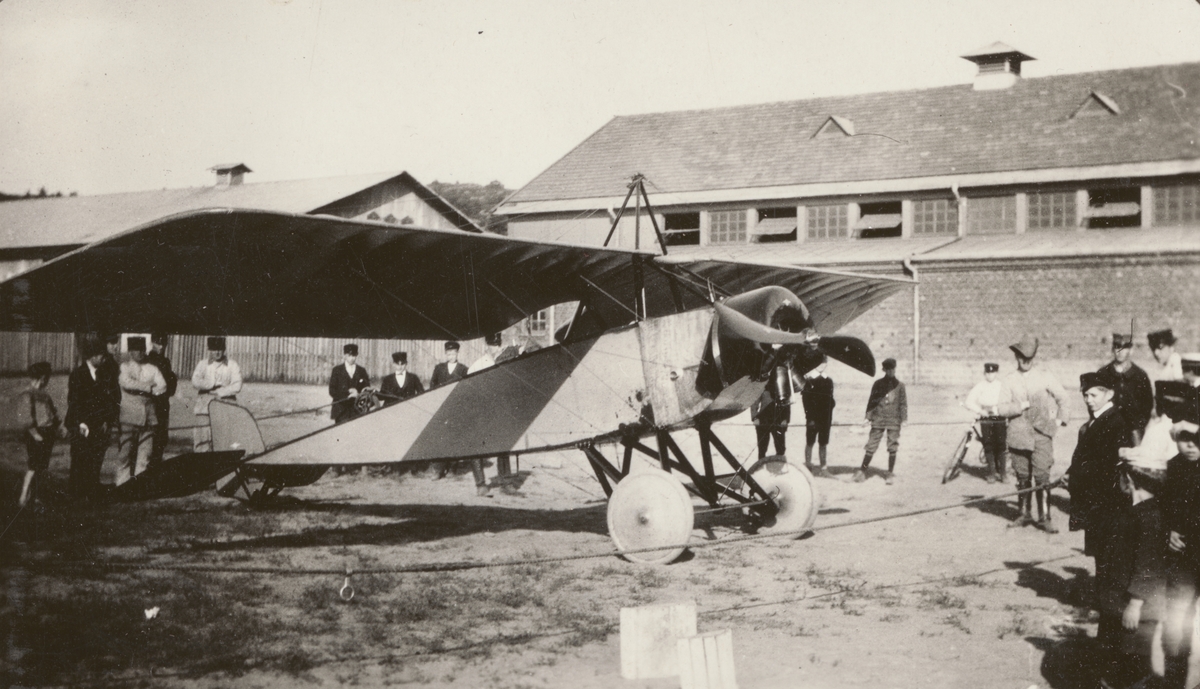 Flygplan Thulin D nummer D.1 visas upp utanför militära byggnader. Folksamling med militärer, civila och barn samlade utanför det inrepade flygplanet, 1915.

Text vid foto i album: "1915"