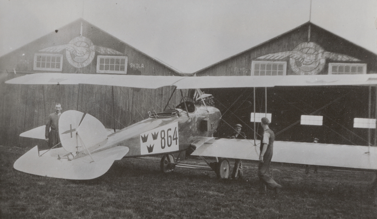 Flygplan Thulin FA nr 864 tillhörande Flygkompaniet på flygfältet vid Enoch Thulins flygskola, cirka 1918-1920. Tre män vid flygplanet. Hangarer i bakgrunden.
