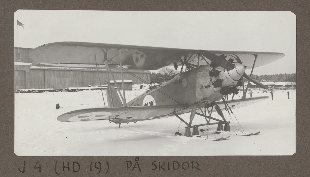 Flygplan J 4, Heinkel HD 19 står med skidor, vintertid, framför hangar på F 2 Hägernäs, cirka 1929-1930.

Text vid foto: "J 4 (HD 19) på skidor."