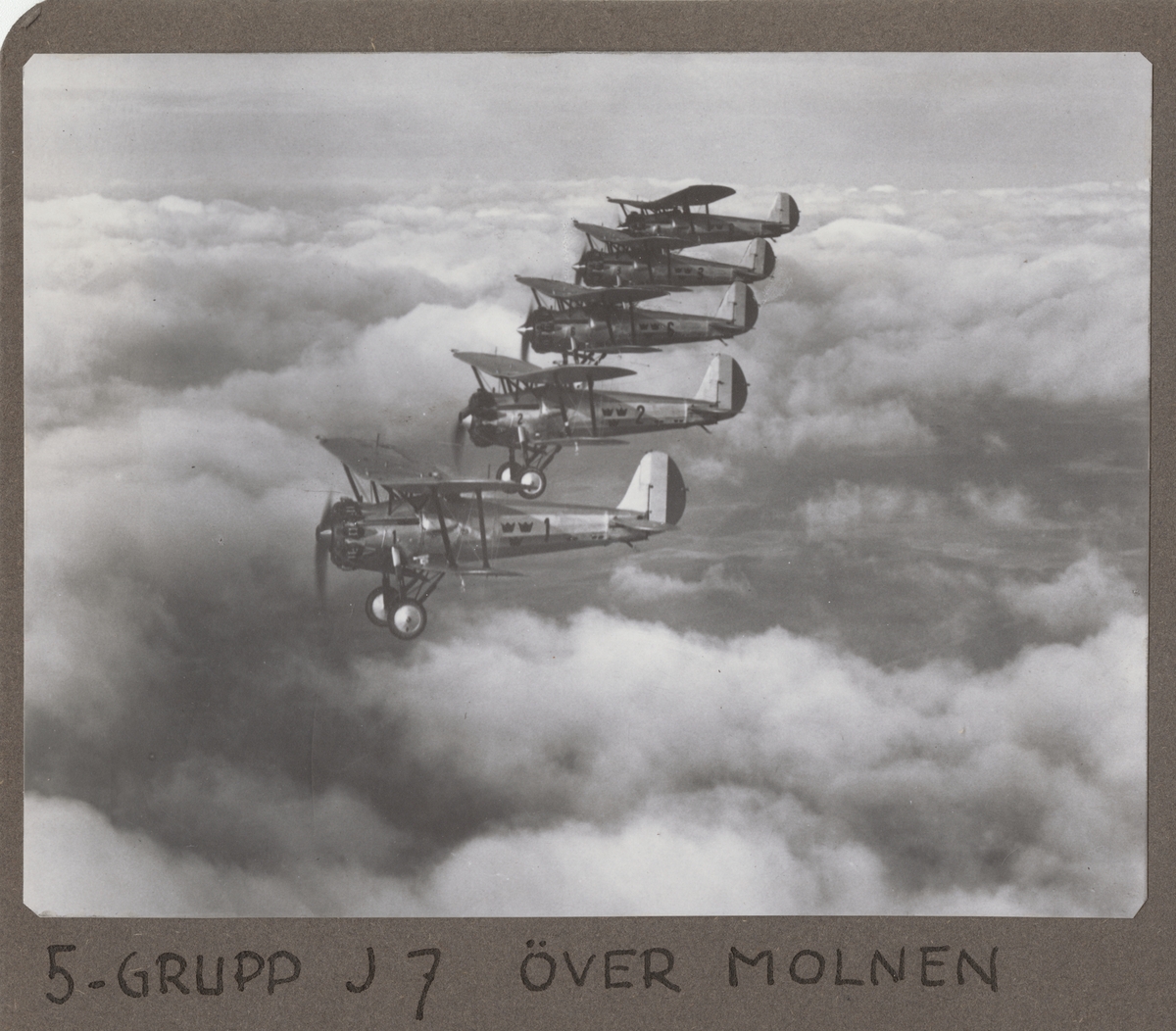 Fem flygplan J 7 Bristol Bulldog flyger i formation ovan moln. Vid Skånemanövern, september 1935. Flygplan från F 1 Västerås, märkta nummer 1, 2, 6, 3 och 8.

Text vid foto: "5-grupp J 7 över molnen."