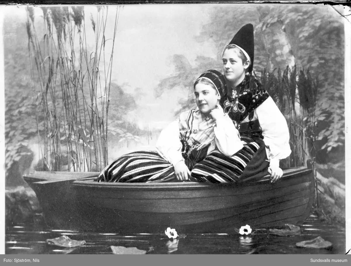 Ateljébild med två unga kvinnor i folkdräkt, sittande i en liten båt.