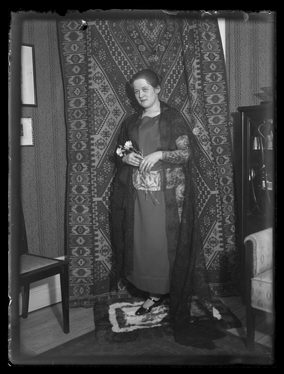 Kvinna i lång klänning och lång schal, stående framför mönstrat draperi. Västerås.