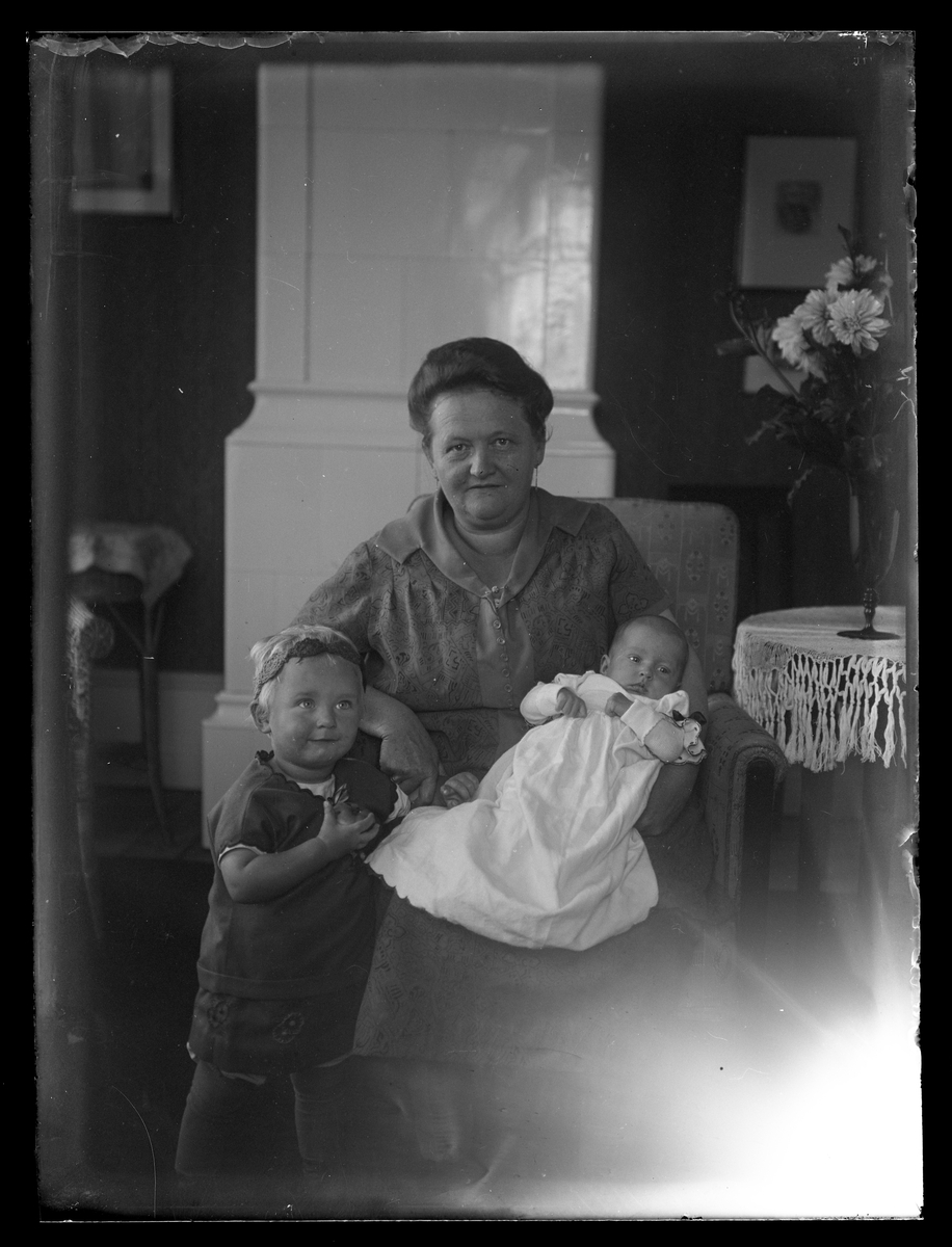 Sittande kvinna med ett barn stående bredvid sig och en baby, klädd i dopklänning, i knät.
Västerås.