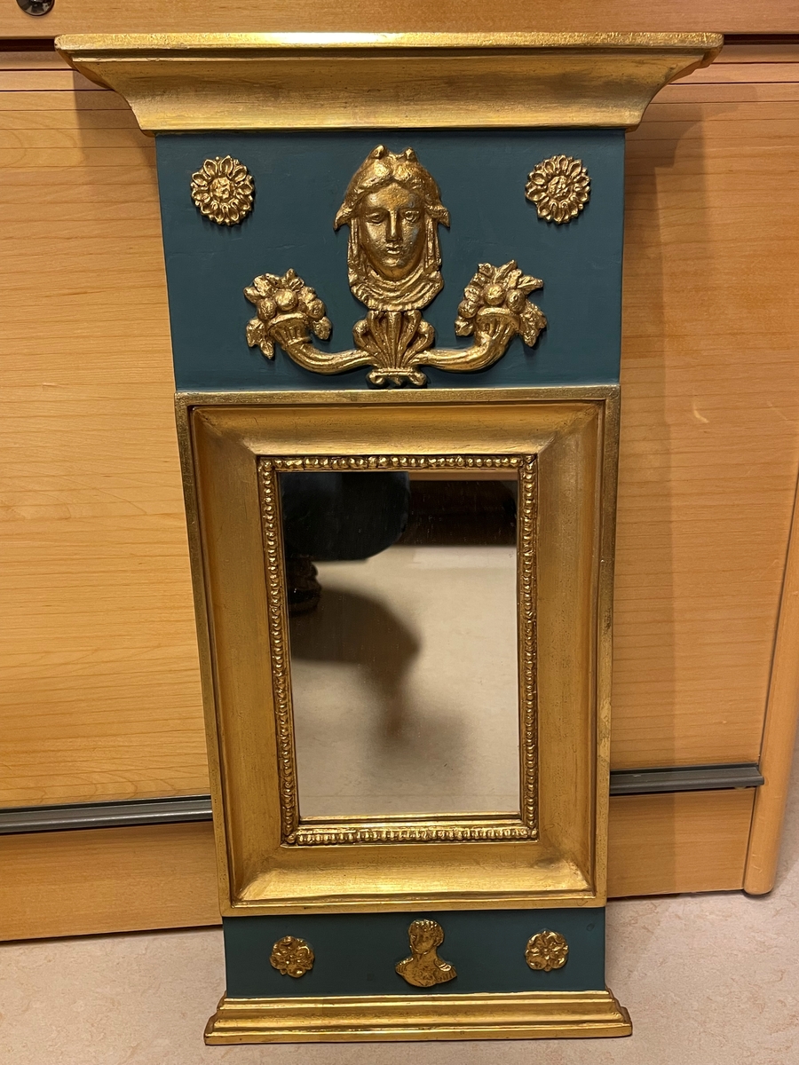 Spegel i empirestil, målad i blått och guld. Reliefmönster med Medusahuvud, ymninghetshorn, solrosliknande blommor, Carl XIV Johans bild och rosor.