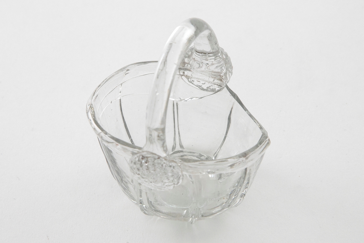 Kurvformet skål i klart glass. Noe sammentrykket korpus som er inndelt med vertikale riller. En bøyd glasstav som fungerer som hank, og overgangen til korpus er markert med to rosetter. Puntemerke på undersiden av skålen.
