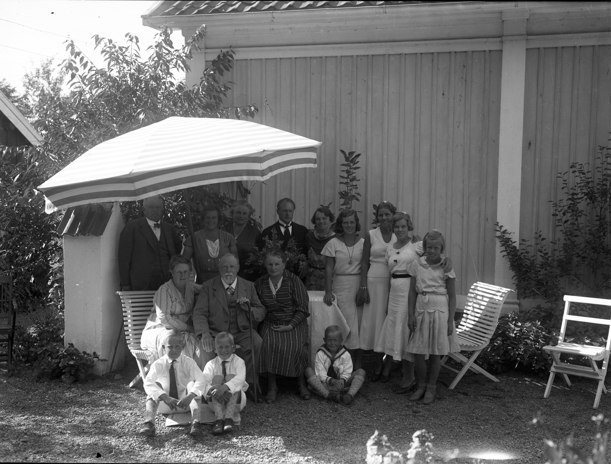 En grupp kvinnor, män och barn står respektive sitter i trädgårdsmöbler under ett parasoll. Det är herr och fru Oldenburg med barn och barnbarn.