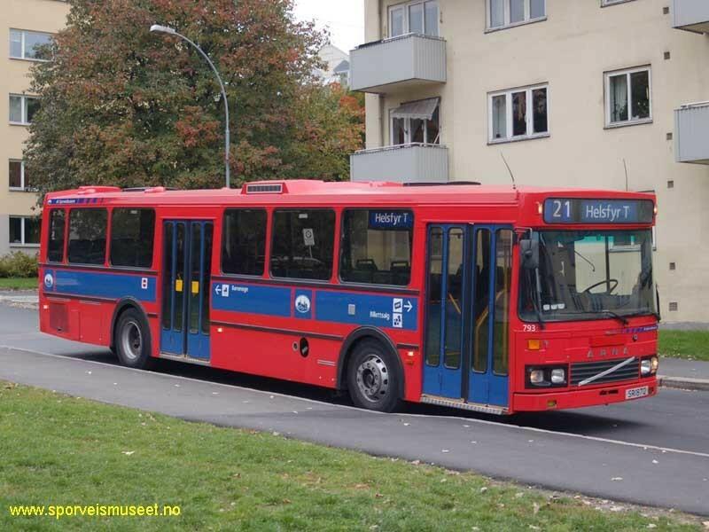 Rød buss med blå dører og et blått bånd på langsiden. Bussen har to dobbeltdører. Bussens interiør består av blå seter, gule stenger og gråe tak og gulv. 