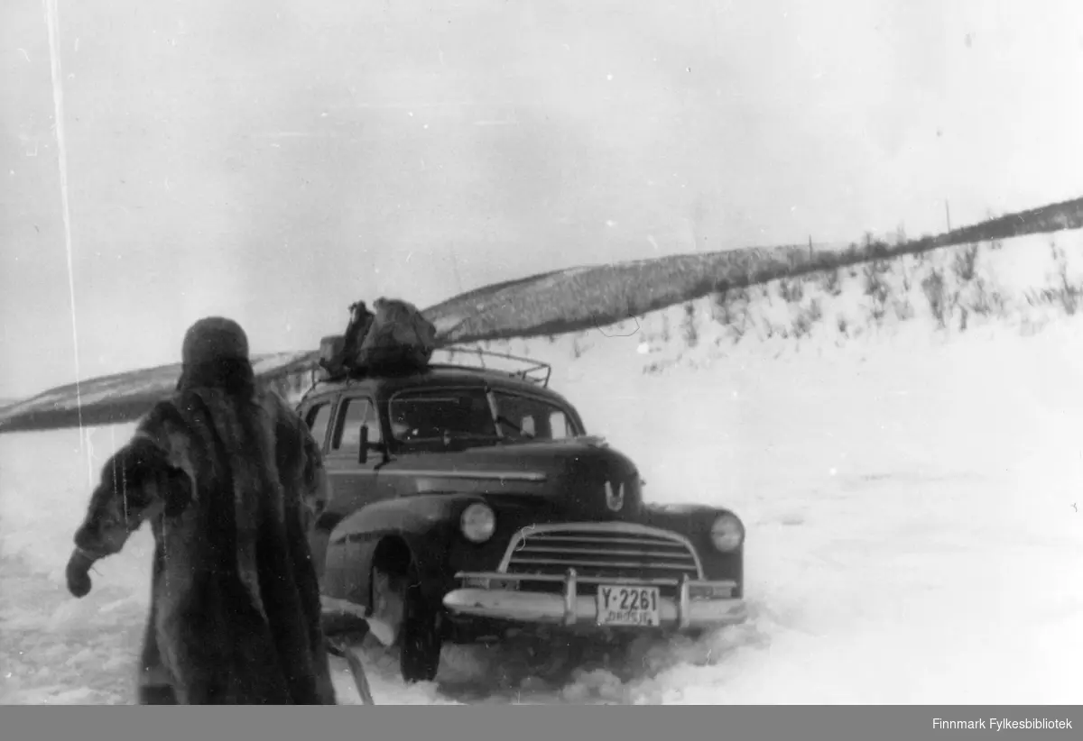 "Det var flere som var uheldige den dagen". 
Drosja, en Chevrolet anno 1946 (Y-2261) står fast på isveien over Tanaelva,  fotografert i tidsrommet 1945-1950. En mann i pelskåpe står ved siden bilen. 