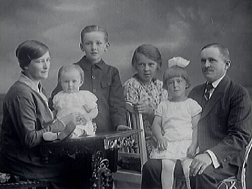 Trävaruhandlare Andersson med familj; hustru och fyra barn. Det minsta barnet sitter på ett bord intill kvinnan. Mannen har en av döttrarna i sitt knä.