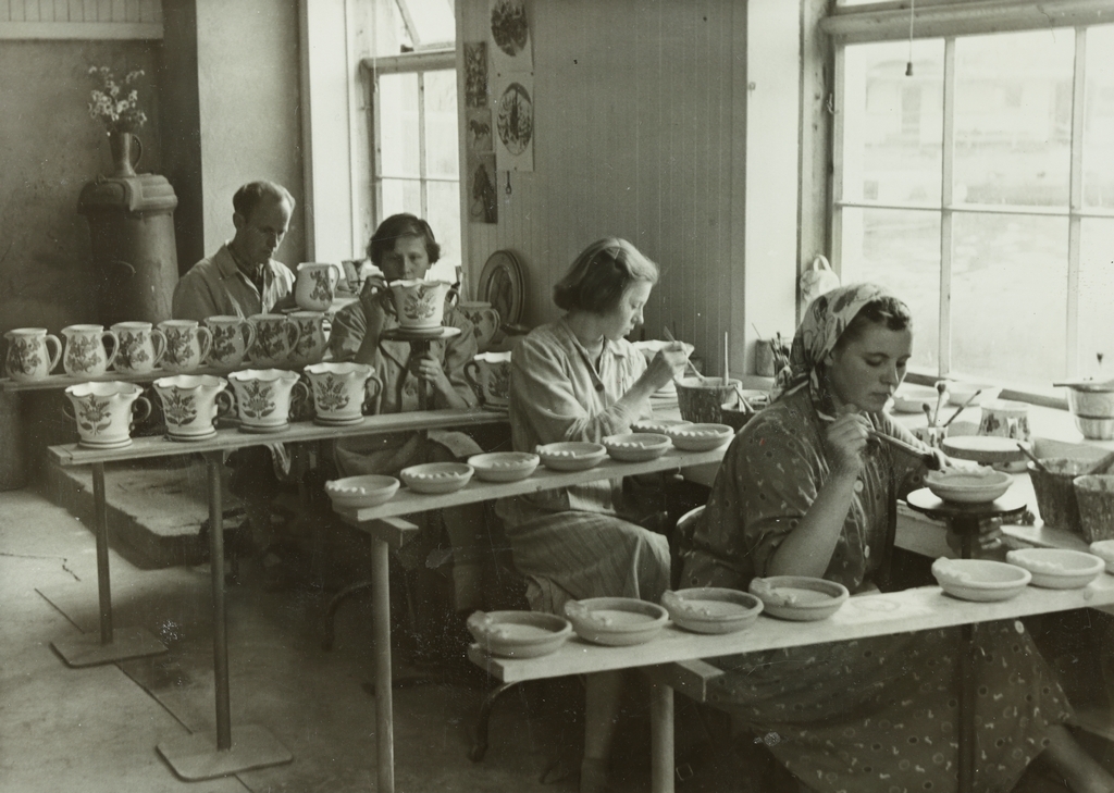 Tre kvinner og en mann sitter og dekorer keramiske produkter som askebegre, mugger og blomsterpotter. Sannsynligvis er fotografiet tatt i produksjonslokalene til Figgjo Fajanse A/S på Figgjo. 