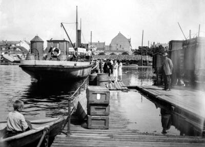 Fotografi av en båt med stavnen mot fotografen, personer som går ombord og en jernbanevogn til høyre i bildet. Strandgata i Hamar skimtes i bakgrunnen.. Foto/Photo