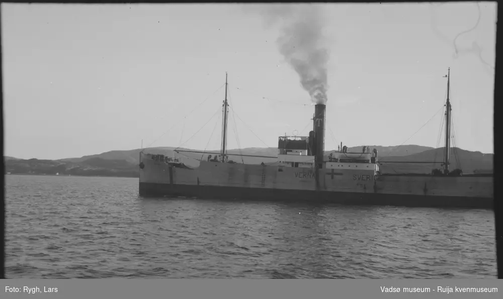 En dampbåt på sjøen, med røyk fra skorsteinen. På skroget står det skrevet VERNA, og SVERIGE. Det er også merket med et flagg. I bakgrunnen er det fjell, og det skimtes noen hus. Bildet har tilhørt Lars Rygh fra Hønefoss. Han var på mineryddeoppdrag i Varangerfjorden, mellom 1917 - 1918.