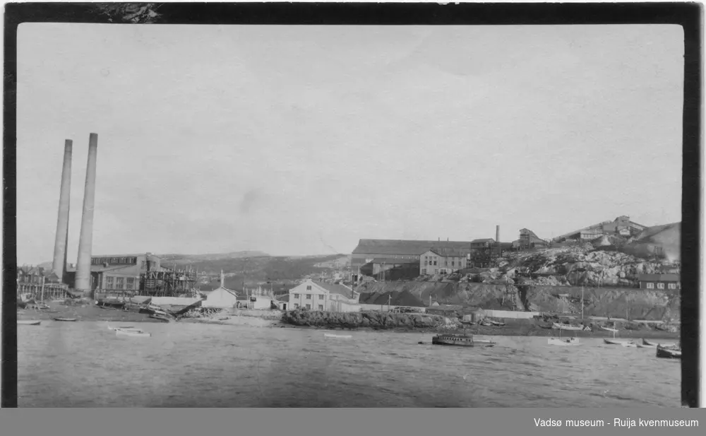 Bilde tatt fra sjøsiden, av Kirkenes havn. Flere av Sydvarangers  anleggsbygninger, og fabrikkpiper kan ses. Det ligger flere trebåter ute på sjøen. Bildet har tilhørt Lars Rygh fra Hønefoss. Han var med på mineryddingsoppdrag i Varangerfjorden 1917 - 1918.
