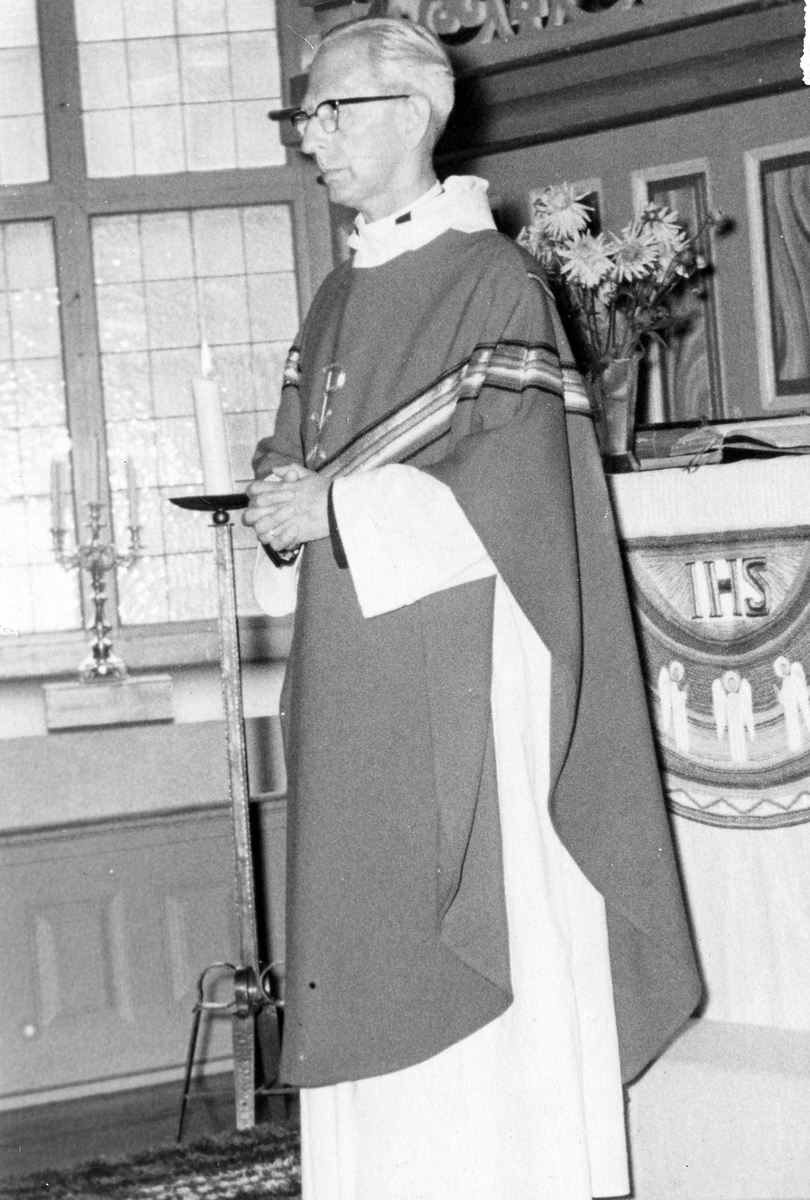 Bild på prosten Karl Linell som står i prästskrud framför ett altare. På altaret hänger ett antependium med text "IHS".