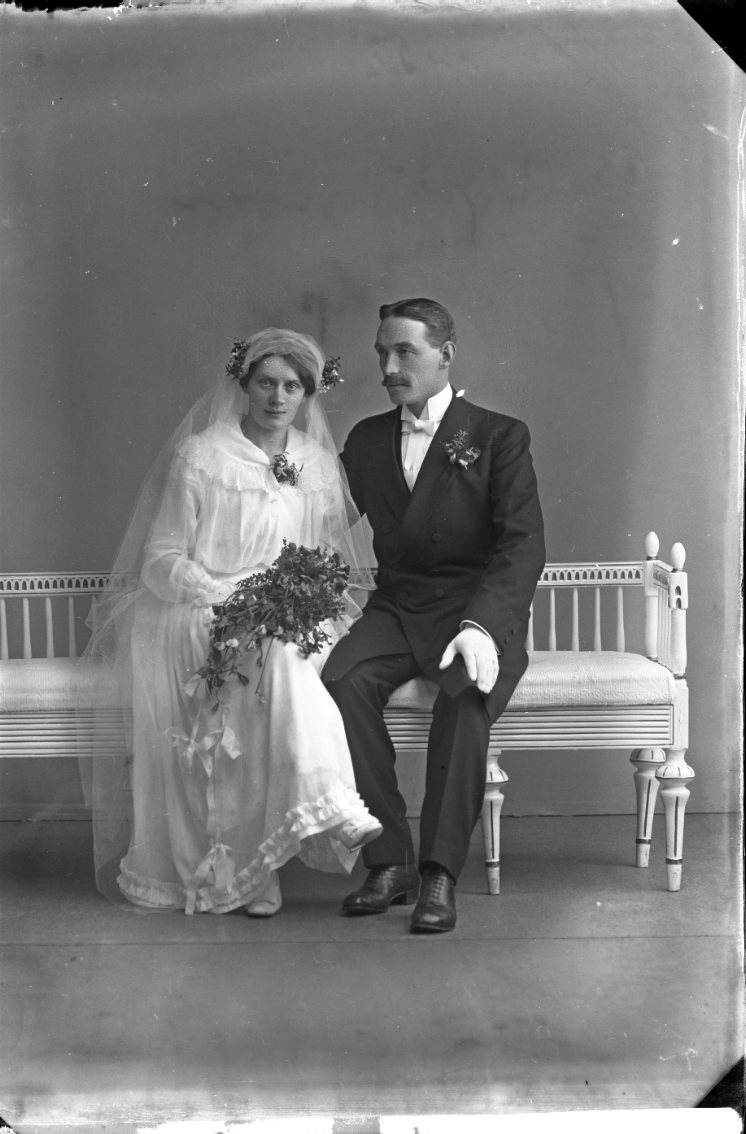 Ett brudpar sitter i en soffa av gustaviansk stil. Bruden är klädd i vitt och håller en bukett i händerna. Brudgummen har vita handskar.