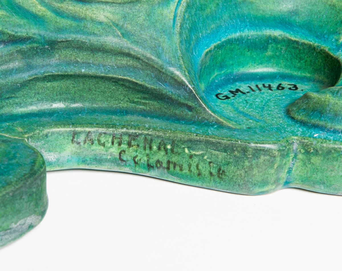 Bordsuppsats i keramik med grön glasyr, formgiven här i lera av den franske keramikern Edmond Lachenal efter en originalskiss i gips av Ida Matton, se XLM.42118. Tillverkad i Paris. Föreställer näcken spelande på harpa vid ett vattenfall.