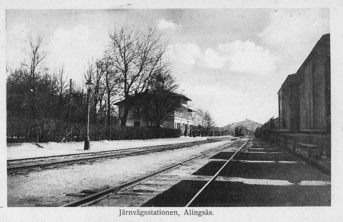 Alingsås Järnvägsstation. Ett tåg är på ingång till väntande människor på perrongen. I fjärran skymtar Härsberget med sitt vattentorn.