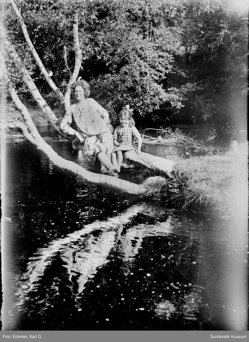 Signe Buckt och Ulla Eckman sitter på en trädstam som hänger ut över Kvarnån i Häre, Indal. Ur Karl Eckmans samling.