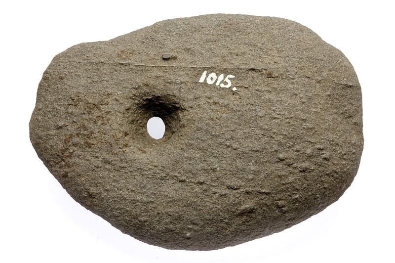 Genomborrad sten, av grå bergart. Oregelbundet oval. Något på sidan om mitten bikoniskt hål.