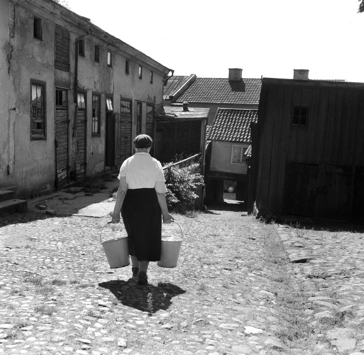 Kvinna som har hämtat vatten i hinkar på en bakgård i Sankt Korskvarteret i Linköping, 1953.
Pressfotografier från 1950-1960-talet. Samtliga bilder är tagna i Östergötland, de flesta i Linköping.