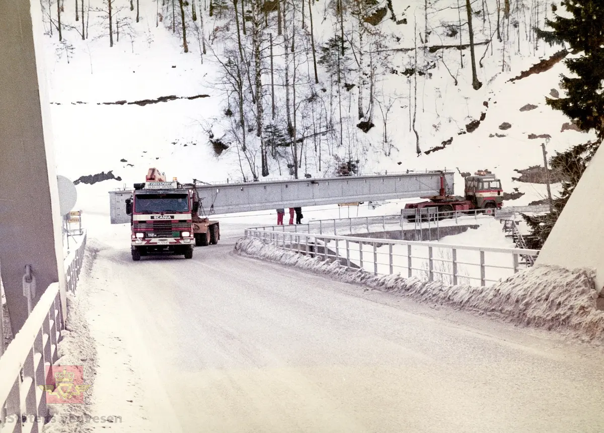 Spesialtransport  av drager til ny Tresnes bru.
Det ble benyttet dragere, hver av dem på 29 tonn.  Her fra transporten av den ene drageren. To  lastebiler ble brukt, så den bakerste kjørte baklengs. Svingene kunne være kritiske.  Dragerene kom med tog til Blakstad jernbanestasjon. Her er transporten i ferd med å kjøre inn på Blakstad bru, som krysser jernbanen og Nidelva.
Lastebil (N3) foran er en 1982 modell Scania med kjennemerke PC51547. Transport Service-Arendal. 
(Kilde: Artikkel om Tresnes bru side 7  i Sørlandsporten Nr.2-1983. Bedriftsavis for statens vegvesen Aust-Agder)
Stedsnavn: Blakstad bru