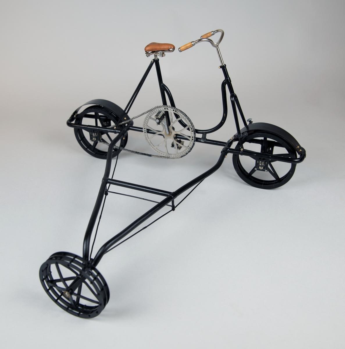 Modell av svart 3-hjulig cykeldressin, skala 1:5, av mässing och stål. Handtag i trä. Kedjan gjord av individuella länkar. Fjädring och broms fungerar. Ljusbrun sadel av formpressat läder.