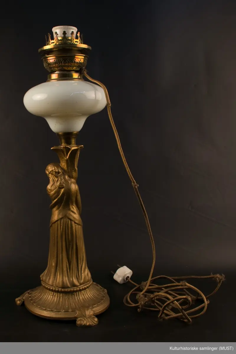 Skulpturell utformet lampefot med kvinnefigur som bærer en fakkel med en kuleformet beholder av hvitt glass.
Opprinnelig parafinlampe. Ombygd til elektrisk i senere tid.
