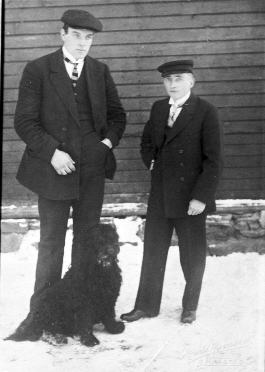 Portrett av to pent kledde menn og en hund stående ved en husvegg.