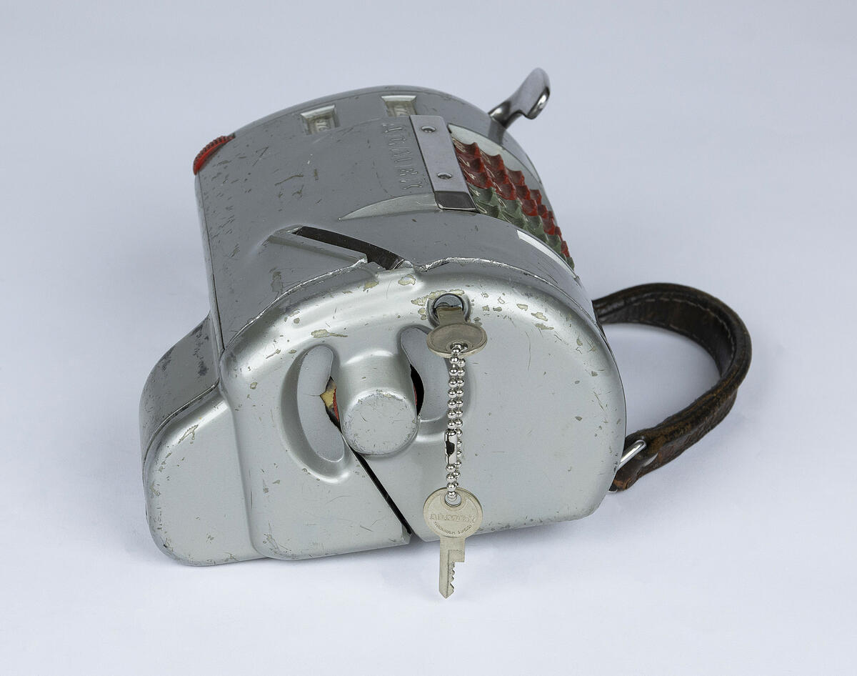 Billettmaskin av typen Almex i metall med en reim i brunt skinn. 