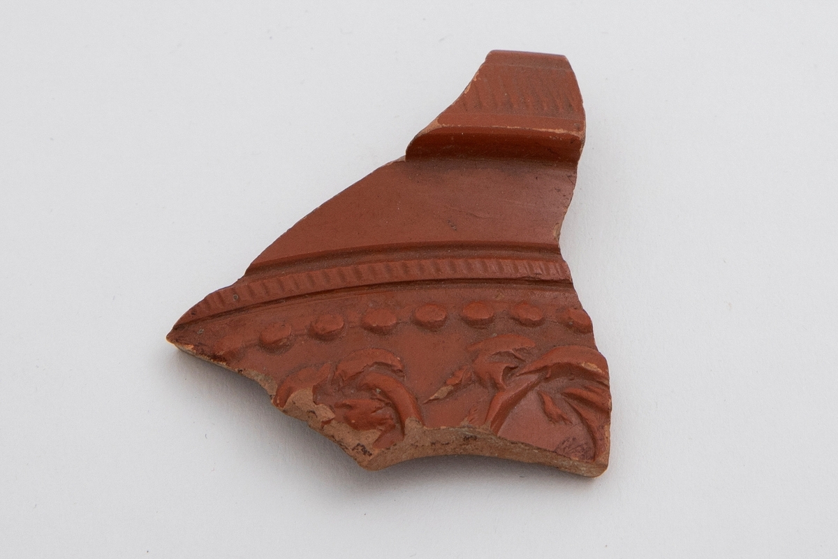 Fragment av arrentinsk vase i terrakotta, dekorert med rødfarget begitning. På yttersiden dekor i lavt relieff i form av en bord med organisk motiv samt profilert kant.