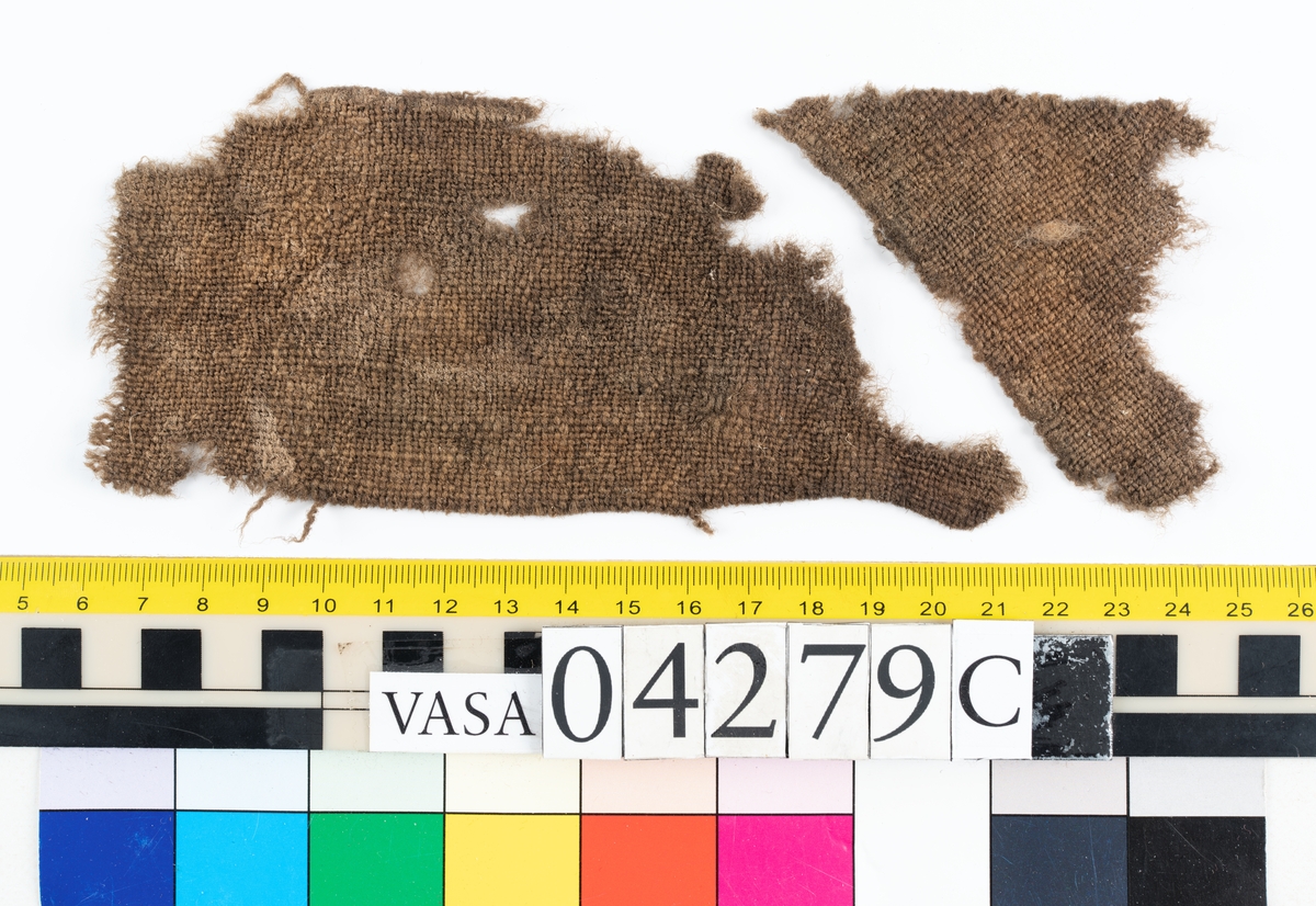 Fyndnumret är fördelat i 16 stycken askar och innehåller runt 350 stycken textilfragment av både ull och bastfiber, eventuellt lin. Tillskärning och sömmar visar att många av fragmenten varit delar av klädesplagg. Exempelvis finns delar av minst en tröja.
.
Textildokumentation 2018.
Textil, fyndnumret består av cirka 349 stycken fragment uppdelade på16 stycken askar (a-p).
Kontextgrupp 18 (inga övriga fyndnummer i gruppen).
Däck: Övre batteridäck.
Relaterade fynd: Inga.
.

Fnr 04279d Typnr 18.2:1-4 
Sammanfattning - Fyra stycken textilfragment av ull vävd i tuskaft. Färgen är nu mörkbrun, originalfärg okänd. Fragmenten är troligen delar av en dräkt.
Textilien har lugg på en sida. Luggen kommer eventuellt från färdigställningsprocessen, i så fall tillkommen genom att tyget är ruggat och överskuret (kläde).
Garnet i trs 1 är något tunnare samt hårdare spunnet än garnet i trs 2. Väven är jämn men garnet varierar något i grovlek. Fragmentens ytor är bitvis matta och har där en mer beige färg än resten av ytan. Detta beror på att den mörkbruna ytan på många av ullfibrerna är bortnött och bara den beige-gula fiberkärnan finns kvar. På ytan i textiliens lugg finns enstaka färgade fibrer.
Andra delar: Inga
Relaterade fynd: Inga.
Fragment 2:1 - Fragmentet har en kant som eventuellt är tillskuren, möjligen har den även stygnhål (söm 1).
Fragment 2:2-4 - Ingen ytterligare information.
.
Fnr 04279e Typnr 18.3:1 
Sammanfattning - Ett fragment av ull vävd i tuskaft. Färgen är nu ljusbrun/mellanbrun, originalfärg okänd. Fragmentet är troligen en del av en dräkt.
Väven är tjock och stabil och därför troligen valkad. Bindningen är bitvis svår att se pga den enhetliga ytan textilien fått då den valkats (eller fått liknande behandling). 
Andra delar: Inga.
Relaterade fynd: Inga. 
Fragment 3:1 – Fragmentet är triangelformat och alla tre kanter är tillskurna. Två av kanterna är något böjda, det beror på att fragmentet troligen är format efter tillskärning och att åtminstone en av dem inte är tillskuren helt rakt från början. Enbart i en av triangelns spetsar saknas eventuellt originalkanter, spetsen ser skadad ut. Fragmentet har inga spår av sömmar.
.

Fnr 04279f Typnr 18.4:1-5 
Sammanfattning - Fem textilfragment av ull vävd i tuskaft. Färgen är nu mellanbrun, originalfärg okänd. Fragmenten är troligen delar av en dräkt.
Textilien är ytbehandlad, troligen ruggad och överskuren (troligen kläde), luggen är mycket tät. Där den är välbevarad går det inte att urskilja vävens bindning.
Andra delar: Inga.
Relaterade fynd: Inga. 
Fragment 4:1 - Fragmentet är som en tidigare konserveringsåtgärd limmat på ett stödtyg, baksidan går därför inte att undersöka. Fragmentet har tre tillskurna kanter. Kant 1 (131 mm lång) har möjligen spår efter en söm (söm 1: Efterstygn). På ytan finns en del bastfibrer samt två stycken, ca 10-13 mm stora, rostfärgade avtryck.
Fragment 4:2-5 - Ingen ytterligare information.
.
Fnr 04279f Typnr 18.5:1-4 
Sammanfattning - Fyra fragment av ull vävd i kypert. Färgen är nu orangebrun/mellanbrun, originalfärg okänd.
Ena sidan har en tydlig ganska lång lugg, medan andra sidan har en kort lugg fläckvis spridd över ytan, eventuellt beror luggen enbart på slitage eller nötning. Inga sömmar finns.
Andra delar: I asken finns en bunt med lösa hår/fibrer (6:1). De låg tillsammans med fragment 5:4, dessa är nu separerade. 18.5 och 18.6 är möjligen samma typ.
Relaterade fynd: Inga.
Fragment ÖB18.5:1-4 – Ingen ytterligare information
.
Fnr 04279f Typnr 18.6:1 
Sammanfattning - Bunt med ull/hår som även innehåller en del bastfibrer. Färgen är nu orangebrun, originalfärg okänd. 
Textilien innehåller två intressanta fibertyper. Den ena karaktäriseras av sin kärna/medulla av typen "uniserial" och kommer möjligen från någon gnagare (Rodentia) eller hardjur (Lagomorpha). Den andra karaktäriseras av sina mycket täta epidermisfjäll och kommer möjligen från samma grupp av djur. Fibrerna kan även komma från samma typ av djur men är då olika typer av hår, dvs underull och täckhår.
Andra delar: Typ 18.6 är möjligen samma typ som 18.5.
Relaterade fynd: Inga.
Fragment 6:1 – Ingen ytterligare information.
.

Fnr 04279g Typnr 18.5:5
Sammanfattning - Ett fragment av ull vävd i kypert. Färgen är nu orangebrun, originalfärg okänd.
Textilien har troligen en ytbehandling, se fnr 04279f typnr 18.5:1-4 för mer information.
Andra delar: Fyndnummer 04279f 18.5:1-4. 18.5 är eventuellt samma typ av textil som 18.6.
Relaterade fynd: Inga.
Fragment 6:1 – Ingen ytterligare information.
.

Fnr 04279g Typnr 18.7:1 
Sammanfattning - Ett fragment av ull vävd i tuskaft. Färgen är nu brun, originalfärg okänd. Fragmentet är troligen en del av en dräkt. 
Textiliens sida A är ytbehandlad, troligen ruggad och överskuren (troligen kläde). Luggen är kort och tät, där den är välbevarad syns inte vävens bindning. 
Möjligen är det samma garn i både varp och inslag. Garnerna är mycket lika både för ögat och i gjorda mätningar, båda är z-spunna.
Andra delar: Inga. 
Relaterade fynd: Ett mycket liknande fragment (tillskärning) finns i en annan ask: 04279i typnummer 18.9:1.
Fragment 7:1 - Fragmentet är i det närmaste ovalt och är tillskuret runt om förutom på en ca 150 mm lång sträcka på ena långsidan och in på en kortsida där kanten är trasig. På den halva av fragmentet där kanten är hel finns både stygnhål och veck/rynkor bevarade längs kanten (söm 1: Sammanfogande). På de partier av kanten där stygnhål finns bevarade är veck/rynkor placerade mellan stygnhålen. Veck/rynkor finns även sporadiskt på partier av kanten där inga stygnhål finns bevarade. Vecken/rynkorna sträcker sig ca 3 mm in från kanten och sitter med ca 6 mm mellanrum. Troligen har en kontinuerlig söm funnits åtminstone längs hela den hela långsidan samt halva de båda kortsidorna, det är troligen sömmen som dragit ihop tyget så veck/rynkor bildats.
.
Fnr 04279h Typnr 18.8:1- ca 60 
Sammanfattning - Cirka 60 fragment av ull vävd i tuskaft. Färgen varierar med nyanser av mörkbrun, gråbrun och mellanbrun, originalfärg okänd. Fragmenten är troligen delar av en dräkt. Samtliga fragment är helt eller delvis täckta med en vaxliknande substans
Andra delar: Inga. 
Relaterade fynd: Inga. 
Fragment 18.8:1 - Fragmentet har en tillskuren kant, skuren på skrådden. Ingen ytterligare information
Fragment 18.8:2 - Fragmentet har en tillskuren kant, skuren på skrådden. Några stygnhål finns bevarade, ur ett av dem sticker det upp en tråd, troligen av bastfiber (söm 1).
Fragment 18.8:3- ca 60 - Ingen ytterligare information.
.

Fnr 04279i Typnr 18.9:1-3 
Sammanfattning - Tre fragment av ull vävd i tuskaft. Samtliga fragment har tillskurna kanter och är troligen delar av en dräkt, ett av dem liknar till sin form ett skört till en tröja. Färgen är brun/mörkbrun, originalfärg okänd. Textilien har rester av en mycket tät och kort lugg på ena sidan. Bindningen är inte synlig där luggen är bevarad. Trådsystem 1 är tätare än trs 2.
Andra delar: Inga. 
Relaterade fynd: Angående fragment 9:1 - ett liknande fragment finns i ask 04279g, typnummer 18.7:1. Formen är mycket lika men de är antagligen tillskurna i olika varpriktning och i olika tyger.
Fragment 9:1 - Fragmentet är tillskuret runt om men har några korta sektioner där kanten är trasig. Ena kortsidans båda "hörn" är så rundade att kortsidan går ihop med långsidorna i en nästan helt rundad form. Den andra kortsidans hörn är skarpare även om också de har en svag rundning. Stygnhål och veck från sömmen finns längs stora delar av kanterna (söm 1: Sammanfogande). Veckens placering och storlek gör att fragmentet ser ut att ha varit sytt med hållning mot något annat. Rester av lugg finns på sida B. Rostfläckar finns på sida A, några av dessa har gått igenom till sida B.
Fragment 9:2 - Fragmentet ser till sin form ut att vara ett skört till en tröja. Alla kanter är tillskurna förutom någon kort sektion där kanten är trasig. Stygnhål och veck från sömmen (söm 1:Sammanfogande) finns bevarade längs kanterna. Rester av lugg finns på sida A. På sida B finns flera rostfläckar, några har en mycket distinkt form. I fragmentet finns ett hål, vid hålets kant finns klumpar med avlagringar och rost (konkretion). På fragmentets yta ligger en 4 mm lång bit av en z-tvinnad tvåtrådig ulltråd.
Fragment 9:3 - Alla kanter är tillskurna förutom någon kort sektion där kanten är trasig. Sporadiska stygnhål finns längs kanten (söm 1: Sammanfogande). Rester av lugg finns på sida A. På sida B finns rostfläckar.
.
Fnr 04279i Typnr 18.10:1- ca 180 
Sammanfattning - Cirka 180 textilfragment av ull vävd i tuskaft. Fragment 1-14 är individuellt numrerade och dokumenterade, dessa har unik information så som tillskurna kanter, stygnhål eller andra formade hål, i vissa fall finns även bevarad sytråd. Färgen är brun/gråbrun, originalfärg okänd. Fragmenten är troligen delar av en dräkt.
På ena sidans yta har nästan alla fragment en ojämt täckande substans. Kan vara rester från tiden på havsbotten men kan också vara original, i så fall eventuellt någon form av vax eller lim. På andra sidans yta finns rester av lugg (kan vara en ytbehandling, ev ruggning och överskärning, som vid tillverkning av kläde).
Garnet i trs 1 är tunnare än i trs 2.
Andra delar: Inga. 
Relaterade fynd: Inga.
Fragment 10:1 - Fragmentet har två tillskurna kanter, båda är invikta mot sida B. Den ena (kant 1) är invikt 5 mm, den andra (kant 2) är invikt 10 mm. Längs båda kanterna finns stygnhål, i ett av stygnhålen finns eventuellt en sytrådsrest (kant 1, söm 1: Sammanfogande), (kant 2, söm 2: Sammanfogande), (kant 2, söm 3: Sammanfogande). Fragmentet har inget spår av den ytsubstans som finns på de övriga fragmenten av samma typ.
Fragment 10:2 - Fragmentet har en tillskuren kant, längs kanten finns stygnhål (söm 1: sammanfogande). Kanten ser ut att ha varit invikt ca 4 mm mot sida A, nu ligger den utvikt. Ytan på sida A har rester av av en mycket nött eventuell lugg och ytan på sida B är sporadiskt täckt av en substans, båda nämnda i sammanfattningen. I fragmentet finns ett format hål som troligen är original. Rester från två till eventuellt ursprungliga hål finns. Fragmentet har ett veck som skulle kunna vara original men det går inte med säkerhet att avgöra.
Fragment 10:3 - Fragmentet har inga originalkanter eller sömmar bevarade. Dock finns ett hål format som en månskära som eventuellt är original. Ytan på sida A har rester av en lugg och sida B är täckt av en nämnd substans, båda nämnda i sammanfattningen..
Fragment 10:4 - Fragmentet har inga originalkanter eller sömmar bevarade. Det finns ett veck som möjligen är original. Ytan på sida A har kort och tät lugg och ytan på sida B är helt täckt av en substans, båda nämnda i sammanfattningen.
Fragment 10:5 - Fragmentet har en tillskuren kant, längs kanten finns stygnhål och sytrådsrester bevarade (söm 1: Förstygn, sammanfogande). Längs kanten finns även några större hål som möjligen är original. Dessa är dåligt bevarade men har en diameter på ca 2-3 mm och sitter (på en del av kanten) med ca 9 mm mellanrum. Ytan på sida B har rester av en substans, nämnd i sammanfattningen. Ytan på sida A saknar lugg, vilket gör fragmentet annorlunda mot resten av fragmenten i samma typnummer.
Fragment 10:6 - Fragmentet har två tillskurna kanter, på den ena finns rester av vad som kan vara ett originalhål. Ytan på sida A har rester av en substans och ytan på sida B har mycket nötta rester av vad som kan varit en lugg, båda nämnda i sammanfattningen.
Fragment 10:7 - Fragmentet har flera kanter som troligen är tillskurna (kant 1,2 och 3), en av dem är tillskuren i en välvd form (kant 1). Tre stygnhål finns bevarade längs kant 2 (söm 1). Kant 1 och 3 har inga sömrester. Ytan på sida A har rester av en substans och ytan på sida B har bevarad lugg,  båda nämnda i sammanfattningen.
Fragment 10:8 - Fragmentet är vikt, om vikningen är original är osäkert. Fragmentet har inga originalkanter, stygnhål eller annan information.
Fragment 10:9 - Fragmentet har en invikt fåll (kant 1) med tydliga märken efter stygn (söm 1: Kaststygn). Stygnen har bara gått igenom ett tyglager. På ena kortsidan av fragmentet finns en tillskuren kant (kant 2), längs den finns flera stygnhål (söm 2). Sida B har en tät lugg, nämnd i sammanfattningen. 
Fragment 10:10-14 - Flera små fragment med stygnhål, ingen bevarad tråd finns. Förekomsten av originalkanter går inte att avgöra.
Fragment 10:15- ca 180 - Ett stort antal mindre fragment utan information. Den övervägande delen har den tidigare nämnda substansen på ena sidans yta. Den andra sidans yta har i flera fall en tät lugg, nämnd sammanfattningen.
. 
Fnr 04279i Typnr 18.11:1-2 
Sammanfattning - Två stycken textilfragment av ull, vävd och valkad. Efterbehandlingen är så kraftig att bindningen inte går att avgöra, ytan är slät möjligen pressad. Fragmenten är troligen delar av en dräkt. Färgen är mörkbrun, originalfärg okänd. 
Båda fragmenten har tillskurna kanter och stygnhål.
Andra delar: Inga. 
Relaterade fynd: Inga. 
Fragment 11:1 - Samtliga kanter är tillskurna (kant 1-5), en del av kant 1 är trasig. Hål efter sömmar finns på en del av kant 1 (söm 1) och längs hela kant 2 (söm 2), inga trådrester finns. På sida A på ytan runt kant 2 finns färgade fiberrester från annat material.
Fragment 11:2 - Alla kanter (kant 1-4) är tillskurna. Kant 1-3 och del av kant 4 är något vikta mot sida B.
Stygnhål finns längs kant 2 (söm 1) och längs kant 4 (söm 2). Dessa stygnhål är sporadiska och svåra att se. Inga trådrester finns.
.
Fnr 04279i Typnr 18.12:1-2 
Sammanfattning - Två stycken fragment av ull vävd och valkad. Den valkade ytan är så tät att bindningen är svår att urskilja, troligen är det en tuskaft. Färgen är mörkbrun, originalfärg okänd. Fragmenten är troligen delar av en dräkt.
Båda fragmenten är tillskurna till smala "remsor" och har stygnhål men ingen bevarad tråd.
Andra delar: Inga. 
Relaterade fynd: Inga. 
Fragment 12:1 - Alla kanter är tillskurna. Ett tydligt stygnhål finns, samt en mängd intrycksmärken/fördjupningar som även de troligen är rester efter stygnhål. En lös tråd finns på ytan. Rester av bastfibrer finns utspritt över ytan.
Fragment 12:2 - En kortsida och en del av en långsida har trasiga kanter, i övrigt är alla kanter tillskurna. Bindningen går till viss del att urskilja och är troligen tuskaft. Rester av bastfibrer finns utspritt över ytan.
.

Fnr 04279i Typnr 18.13:1 
Sammanfattning - Ett textilfragment av ull vävd i tuskaft. Färgen är brun, originalfärg okänd.
Fragmentet är en stadkant, inslagsgarnet (trs 2) vänder i ena långsidan. Varpgarnet (trs 1) är tvåtrådigt och mycket grövre än det entrådiga inslagsgarnet.   
Längs med fragmentet finns flera ovala hål, ca 1-3 mm i diameter, samt flera stygnhål.
Andra delar: Inga.
Relaterade fynd: Inga. 
Fragment 13.1 – Ingen ytterligare information.
.

Fnr 04279j Typnr 18.14:1-2 
Sammanfattning - Två stycken fragment av ull vävd i tuskaft. Färgen är orangebrun, originalfärg okänd. Fragmenten är troligen delar av en dräkt. Tyget är kompakt och ganska tjockt, möjligen valkat (stampat).
Ytan på sida B är täckt av en jämn oskuren lugg. Ytan på sida A har en sporadisk lugg, om den är original eller kommer av nötning är osäkert. Textilien är bitvis täckt av salt och rost, samt har rester av en vaxliknande substans. Om substansen är original eller en förorening från tiden på havsbotten är oklart.
Andra delar: Inga.
Relaterade fynd: Inga. 
Fragment 14:1 - En av fragmentets kanter kan möjligen vara en rest av en originalkant med söm (söm 1). Fragmentet har bågformade veck särskilt koncentrerade runt ett hörn, vecken blir mindre bågformade ju längre från hörnet de sitter. I några av vecken har bristningar i tyget uppstått. Ytan på sida B är nästan helt täckt av en tät lugg, ytan på sida A har fläckvis bevarad lugg, särskilt välbevarad i vecken. På ytan på sida A finns spridda bastfibrer, samt på ett ställe rosa fibrer.
Fragment 14:2 - Fragmentet har en 27 mm bred stadkant längs ena sidan. Övriga kanter är troligen tillskurna. Längs stadkanten finns veck som eventuellt kan vara rester av en söm. Över hela ytan finns veck/rynkor. Ytan på sida B är nästan helt täckt av en tät lugg. Ytan på sida A har sporadisk lugg, särskilt i vecken. Mitt på fragmentet finns ett 40 mm långt hål/genomgående skåra, troligen ursprungligt.
.
Fnr 04279k Typnr 18.15:1-2 
Sammanfattning - Två fragment av ull vävd i tuskaft. Färgen är brun,  originalfärg okänd. Tyget har en mycket tät och fin lugg på båda sidor, där den är välbevarad syns inte vävens bindning alls. Garnet i båda trådsystemen är mycket jämnt vilket ger väven en mycket tät och jämn struktur.
Andra delar: Inga.
Relaterade fynd: Liknande delar är: 09421, 09316, 07732 samt 13136. Dessa har suttit ihopmonterade till en mössa (gjort i modern tid för utställning).
Fragment 15:1 - Fragmentets alla kanter är tillskurna och har spår av sömmar i form av stygnhål, intrycksmärken, veck och trådrester. Löst liggande på ytan finns en rest av en tråd. Den är svart, och kommer eventuellt från montering till utställning. Även sömmarna kan komma från samma montering.
Fragment 15:2 - Fragmentets alla kanter är tillskurna och har spår av sömmar i form av stygnhål, intrycksmärken, veck och trådrester. Sömmarna kan komma från montering till utställning.
.
Fnr 04279n Typnr 18.17:1- ca 50 
Sammanfattning - 50 stycken fragment stora nog att räkna individuellt, samt flera mindre fragment, trådar och fibrer från samma textil. Fragmenten består av bastfiber vävd i kypert.
Textilien har varit tvåfärgad. Trs 1 består av 2 olika garn, till övervägande del garn 1 som är gråbrunt och mörkare än det sporadiskt förekommande garn 2 som är beigt. Trs 2 består av garn 3 som är beigt. Garn 2 och 3 är eventuellt samma garn. Pga färgskillnaden i garnerna får textilien en melerad färgeffekt. 
Samtliga garn är ojämnt spunna men textiliens skick och nedbrytningsgrad gör uppskattning och mätningar svåra. Textilien ligger i flera lager vilket gör att fragmenten är upp till 12 mm tjocka.
Inga rester av tillskärning, originalkanter, eller sömmar finns. Textilien är starkt nedbruten, impregnerad av rost och eventuellt delvis mineraliserad.
Andra delar: Inga.
Relaterade fynd: Inga. 
Fragment 1- ca 50 – Ingen ytterligare information.
.
 Fnr 04279o Typnr 18.18.1-2
Sammanfattning - Två fragment av ull vävd i tuskaft. Färgen är orangebrun, originalfärg okänd. Tyget har hårig lugg på båda sidor, det är dock svårt att avgöra om den är original eller uppkommit genom  nötning på textilien. Väven är tät och jämn.
Andra delar: Kan vara samma typ som 04279f 18.6:1.
Relaterade fynd: Inga. 
Fragment 18:1 - En del av en kant är eventuellt tillskuren, där finns ett hål som eventuellt är ett stygnhål. På ytan av både sida A och B finns av en vaxartad beläggning som bygger upp till vaxartade klumpar, beläggningen är troligen ej original. På sida B finns 2 cirkelformade avtryck, de är rostfärgade med vit kant. Fragmentet har varit vikt och någonting som legat emellan har gjort avtrycket.
.
Fnr 04279p Typnr 18.19:1- ca 26 
Sammanfattning - 26 stycken textilfragment (plus fibrer och mindre fragment som inte går att räkna individuellt) gjorda av ull vävd i tuskaft. Textilien är impregnerad av rost och därför orangebrun, originalfärg är okänd. 
Några av fragmenten kan vara delar av axelkarmar.
Sida A har lugg på delar av ytan men troligen har det uppkommit då textilien nötts snarare än att vara en ytbehandling. Flera av fragmenten har tillskurna kanter och stygnhål.
Flera av fragmenten har vaxartade avlagringar på sida A, dessa är upp till några mm tjocka. Textilien är starkt påverkad, impregnerad och missfärgad av rost.
Andra delar: Inga.
Relaterade fynd: Eventuellt 10950.
Fragment 19:1 - Fragmentet har två tillskurna kanter, båda mycket trasiga. En tredje kant är bitvis vikt mot sida B, om vikningen är original är osäkert. De tillskurna kanterna har bevarade stygnhål vilka är sporadiska och otydliga (söm 1 och söm 2). Fragmentets sida A har en stor mängd vaxliknande avlagringar samt en del utspridda bastfibrer.
Fragment 19:2 - Fragmentet har inga tydliga tillskurna kanter men eventuellt är en skåra i fragmentet original. Enstaka stygnhål finns bevarade, minst två av dem har rest av tråd i hålet (söm 1). Sida A har en stor mängd vaxartad avlagring, sida B endast en liten mängd av densamma.
Fragment 19:3 - Fragmentet har minst en, möjligen fler, tillskurna kanter. På båda sidor finns rester av bastfibrer.
Fragment 19:4 - Fragmentet har en tillskuren kant med stygnhål och rester av tråd (söm 1).
Fragment 19:5 - Fragmentet är vikt sida B mot sida B längs trs 2, en långsida utgörs av vikningen. En av kortsidorna är tillskuren och där finns hål i storleken som stygnhål. Om det är stygnhål är dock inte säkert, några av dem ser mer stansade ut vilket gör att de kan ha annat ursprung, de kan eventuellt vara från skadedjur. Hålen går genom båda lagren med tyg.
Fragment 19:6 - Fragmentet har två vikmärken och en tillskuren kant. Längs den tillskurna kanten finns stygnhål med trådrester bevarat i hålen (söm 1). Om fragmentet viks ihop längs den skarpa mittersta viklinjen, nästan på skrådden, sida A mot sida A, möts stygnhålen. Fragmentet har troligtvis varit monterat vikt i original.
Fragment 19:7 - Fragmentet kan vara en del av en axelkarm. Fragmentet har en viklinje ungefär längs trs 2. Det har varit vikt sida A mot sida A, vikningen kan vara original. Fragmentet har en rundad kant som möjligen är tillskuren, stygnhål finns längs kanten (söm 1). Stygnhål finns även en bit in från fragmentets kanter, hur dessa hänger ihop i en söm är otydligt (söm 2).
Fragment 19:8-9 - Fragmenten kan vara delar av en axelkarm. Fragment 8 är vikt ungefär längs trs 2 och har troligen suttit ihop med fragment 9, också vikt ungefär längs trs 2. Sammanlagd längd är ca 170 mm. Vikningen är troligen original. På fragment 9 följer inte hela vikningen trs 2 utan viker av och går sedan mer på skrådden. Längs den ovikta kanten på fragment 9 finns rester av en söm (söm 1). På fragmentet finns 2 övriga stygnhål.
Fragment 19:10 - Fragmentet är vikt längs trs 2 och något snett jämfört med trådsystemet liksom fragment 9, vikningen kan vara original. I övrigt är fragmentet trasigt runt alla kanter. 
Fragment 19:1- ca 26 - Flera mindre fragment utan ytterligare information. Flera av dem består av tyg i flera lager med vikt kant (liksom flera av de numrerade fragmenten ser ut). Fragmenten är genomdränkta av rost och har även den vaxartiga substansen som beskrivits ovan.
