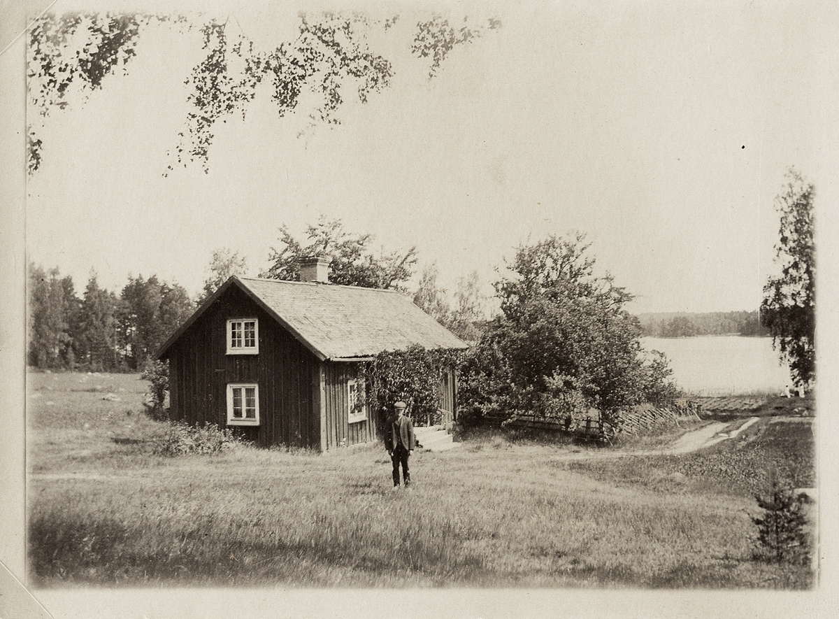 Ett torp någonstans i Kronobergs län. En man i keps poserar framför huset.
I bakgrunden skymtar några kvinnor vid sjön. 1910-tal.