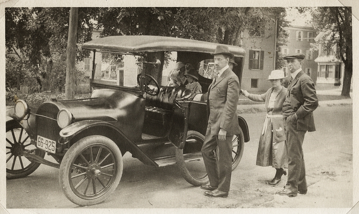På långtur. Två män och en kvinna poserar utanför en bil (efter däckbyte?). I bakgrunden syns några bostadshus.
(USA?). 1920-tal.