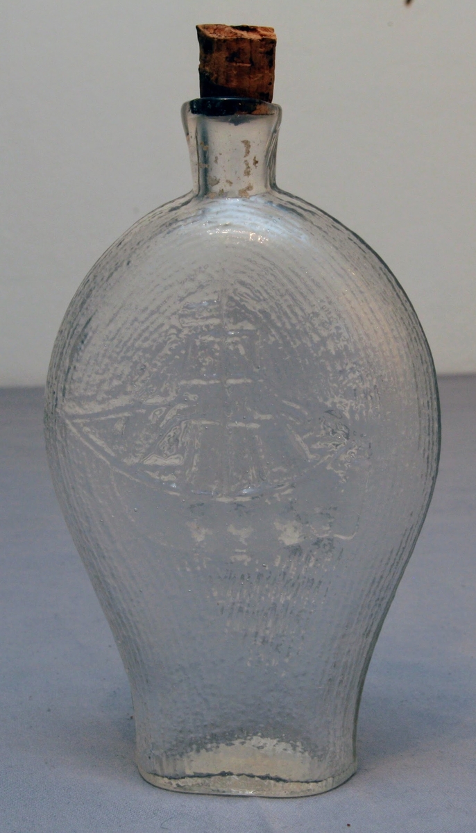 Glasflaska med plan botten och uppåt vidgad form. Med reliefdekor föreställande smala ränder som täcker flaskans ytor. På flaskans ena sidan är ett skepp avbildat, på den andra en druvklase. Med kork.