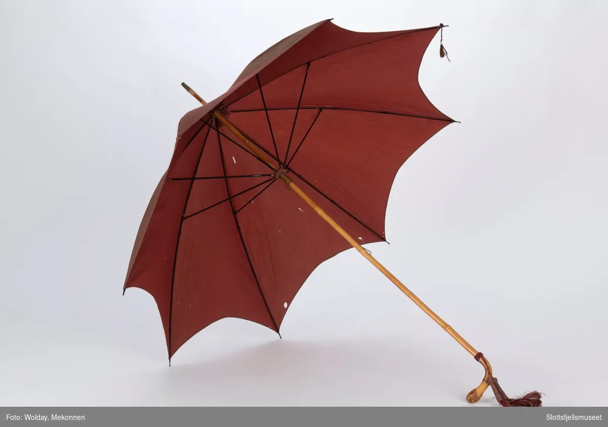 Paraply med 8 spiler av metall og skaft av bambus. Trukket med rødbrun stoff. To dusker festet til skaftet. 