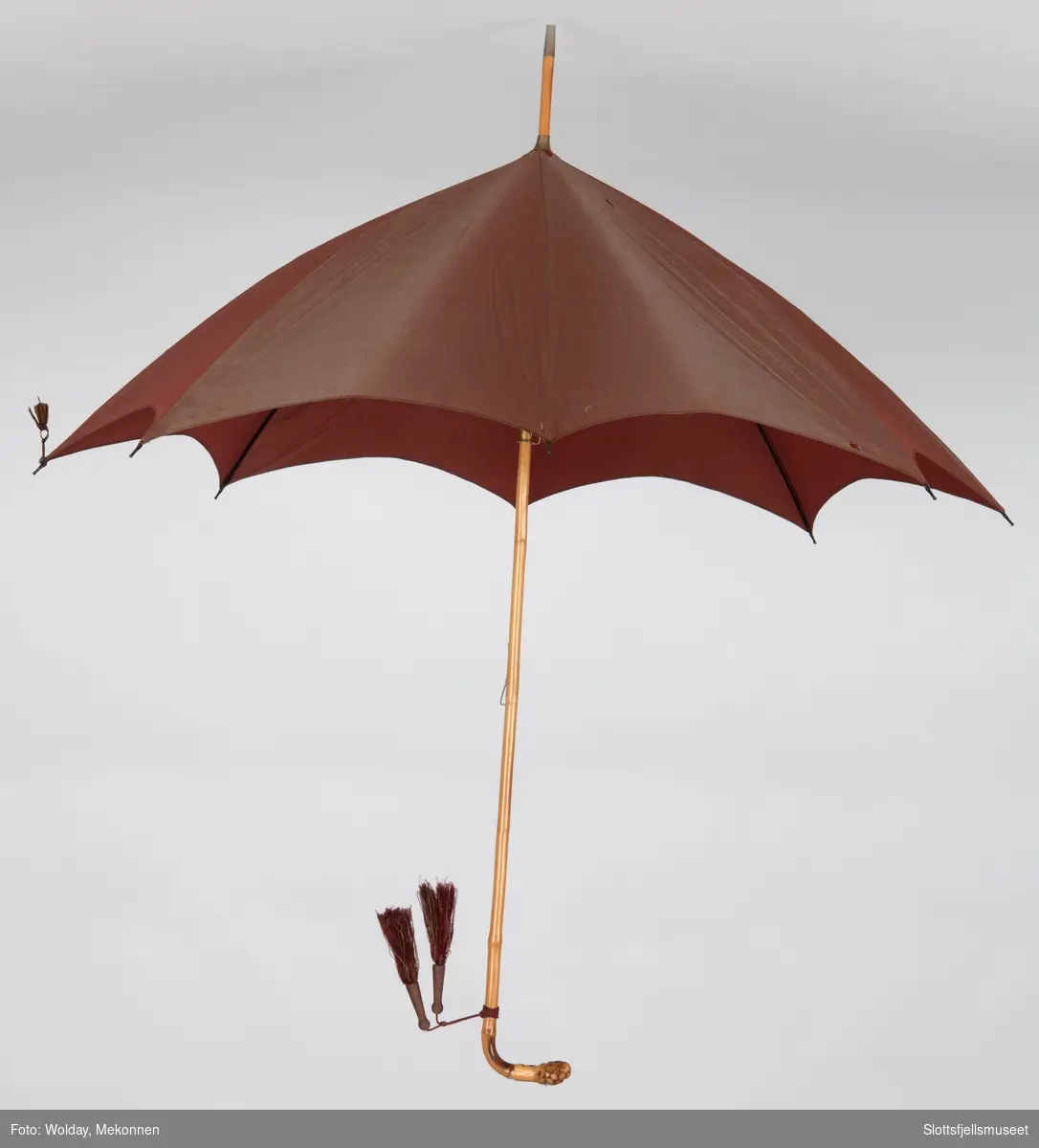 Paraply med 8 spiler av metall og skaft av bambus. Trukket med rødbrun stoff. To dusker festet til skaftet. 
