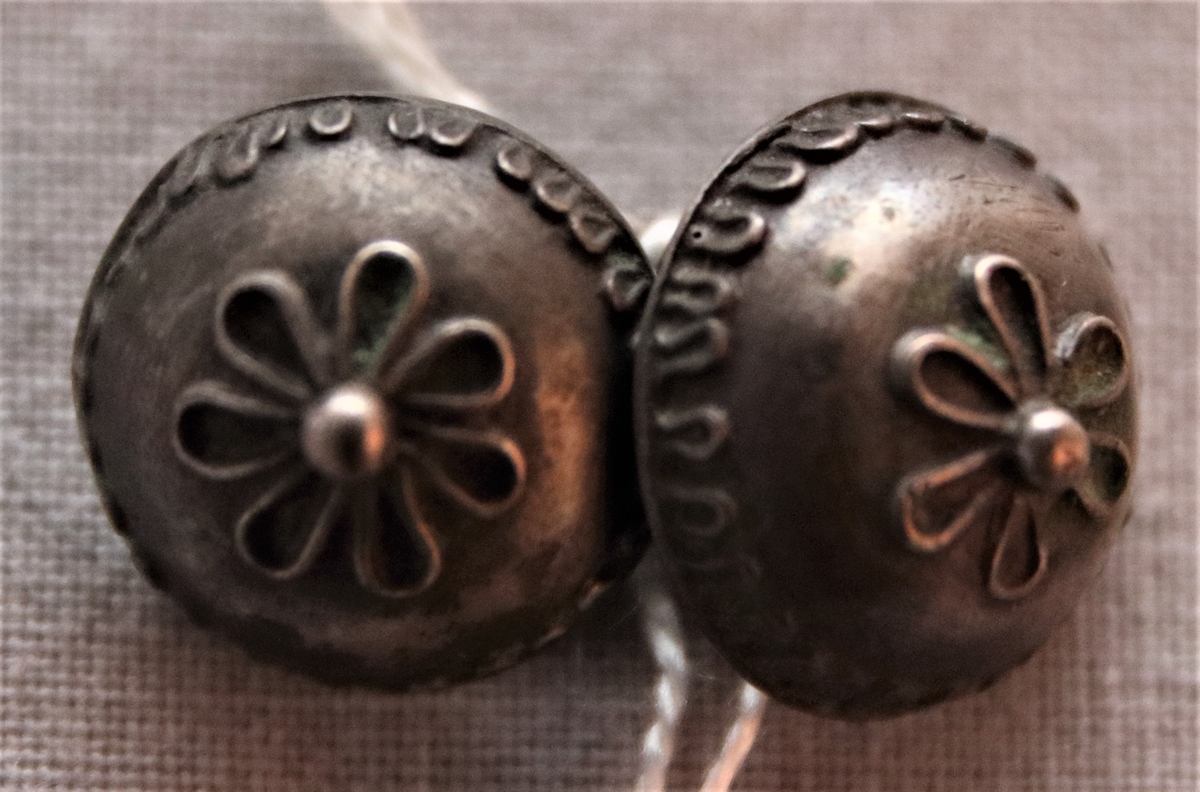 Två buckleformade knappar av tenn, vars märlor sammanlänkats av en mindre ring. Dekor i relief föreställande blommor i mitten av knapparna och en bård längs kanterna.