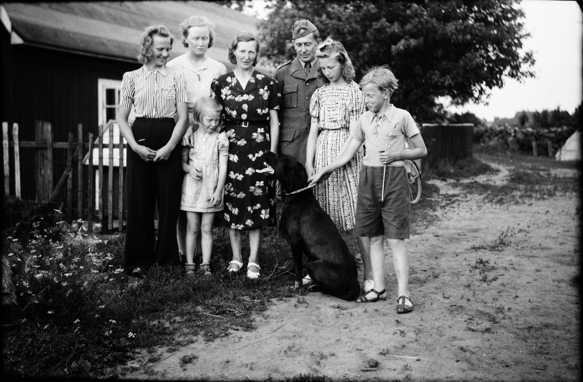 En man i uniform står utanför en gård tillsammans med kvinnor och barn samt en svart hund som sitter och tittar upp på kvinnan i mitten. Alla betraktar hunden utom hon som möter kameran.
(Se även bild EA1517)