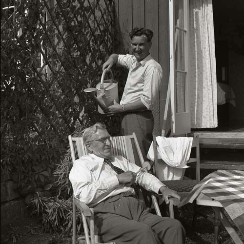 Två män, Jon och Effe, utomhus vid en trädgårdsmöbel i Hamar, Norge 1 juli 1956. Den ene mannen står bakom den andre, han ler, och låtsas hälla ur en vattenkanna över den andres huvud.