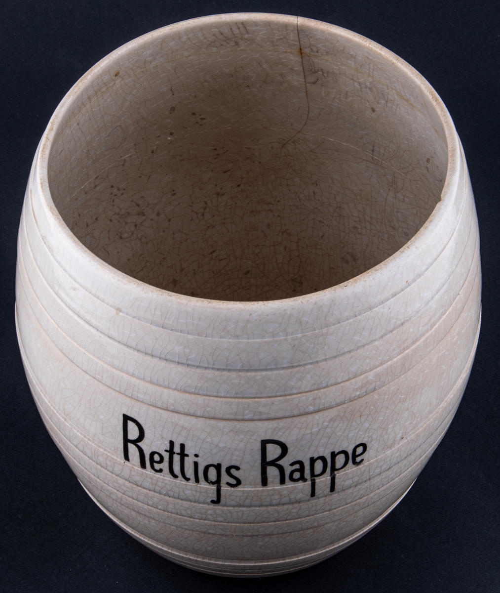 Burk av vit keramik, krackelerad glasyr. Lock saknas.
Text på framsidan: Rettigs Rappe.
Undersidan märkning i godset: 6.