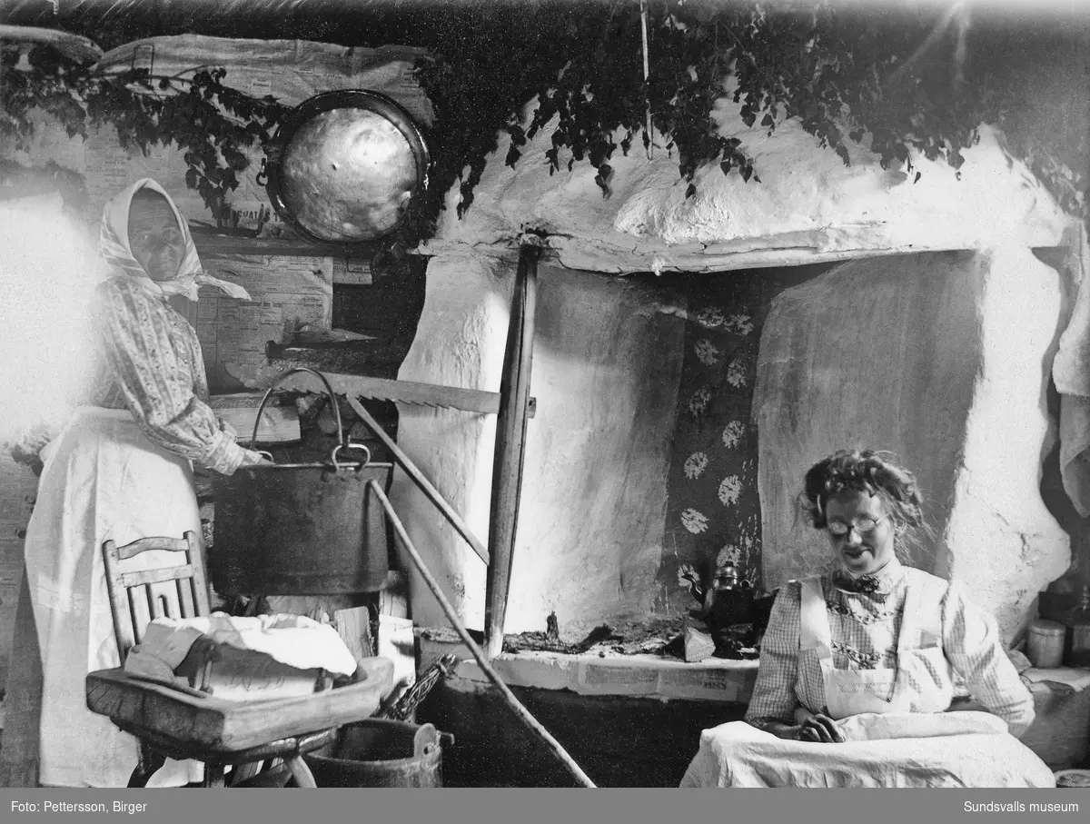 Två äldre kvinnor i ett fäbodkök med öppen härd och lövad spiskåpa i Klackbodarna, Stöde.